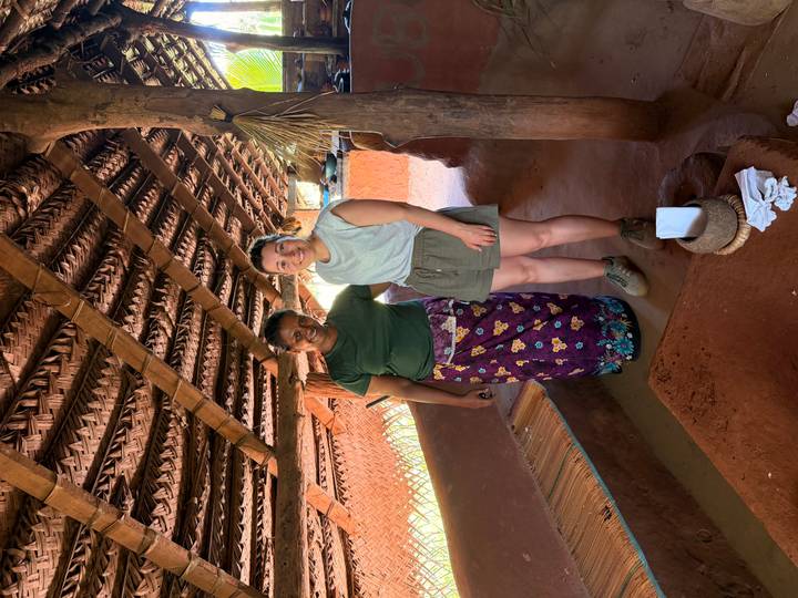 Visitor stands with a local woman inside a rustic thatched kitchen hut.