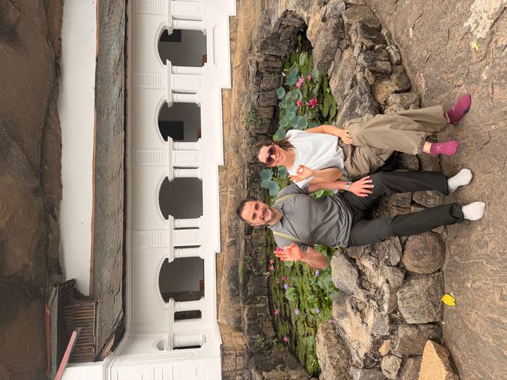 Travellers seated barefoot beside a lotus pond at a white temple built into rock.