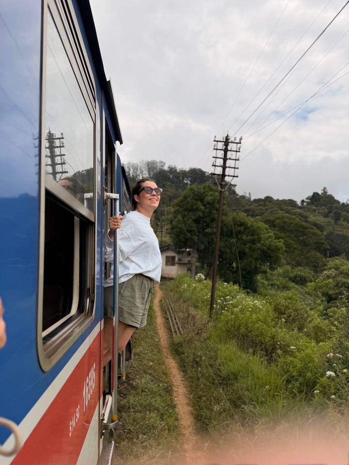 Woman leans out of a blue train enjoying views of misty green highlands.