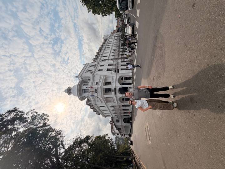 Couple stands on a street corner in front of a grand colonial hotel under a patterned sky.