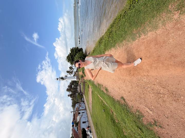 Woman poses on a coastal path with the white Galle lighthouse and sea behind.