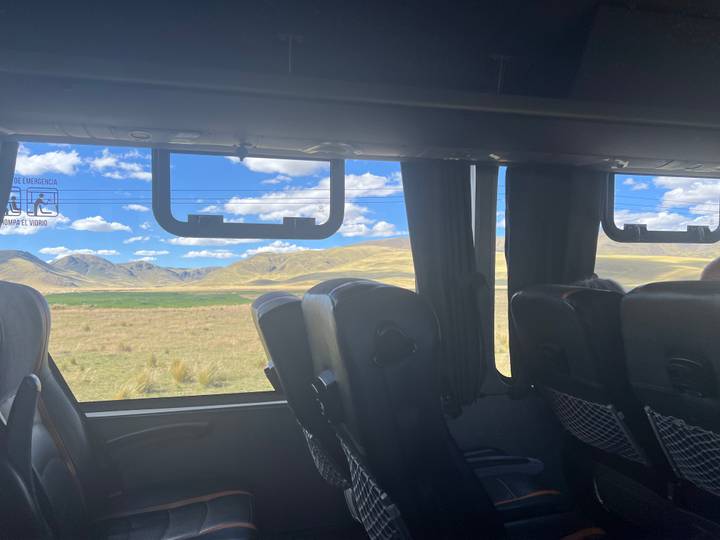 View from inside a tour bus showing empty seats and wide windows framing rolling yellow Andean hills
