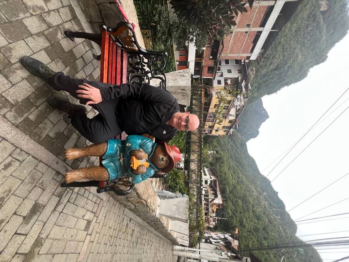 Tourist sitting on a bench beside a Paddington bear statue with lush mountain village backdrop