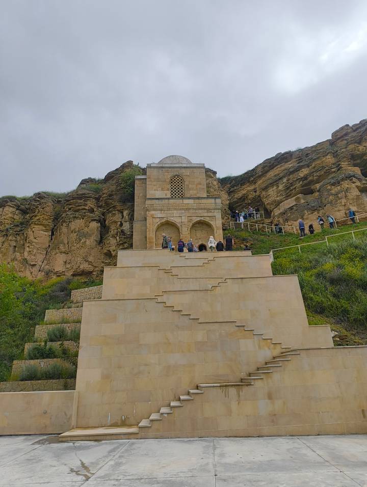 Tourists climbing wide stone steps toward a small mausoleum built into a cliffside of layered rock.