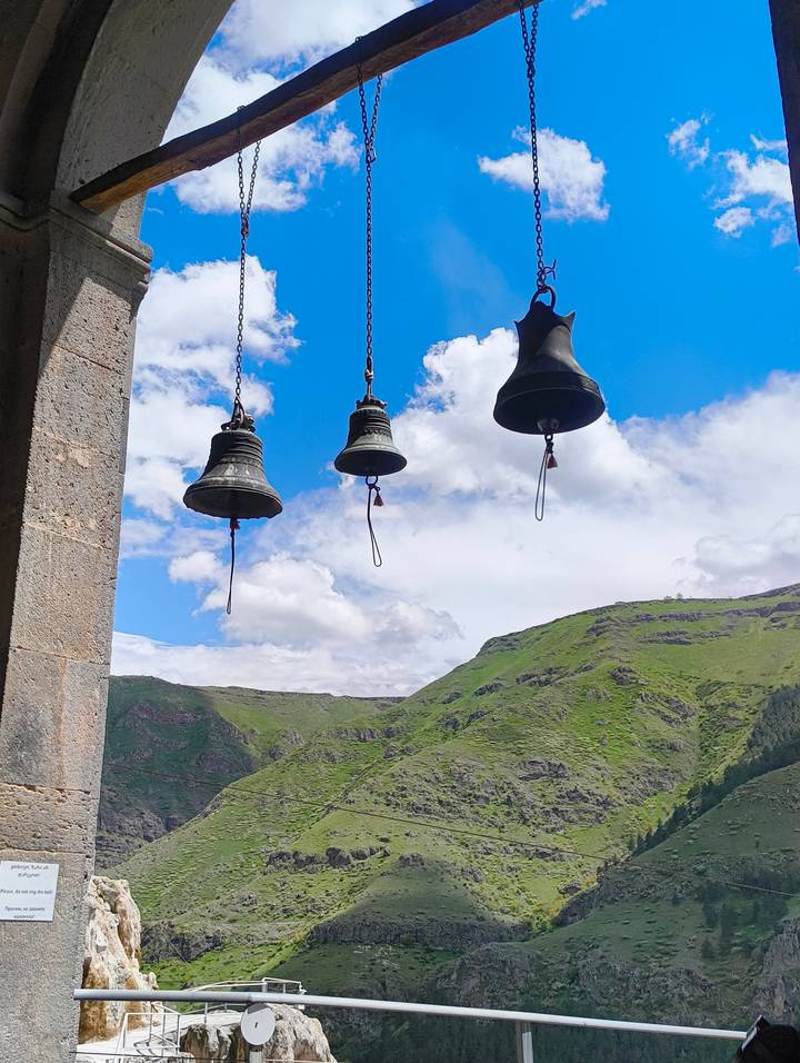 Three church bells hanging in silhouette against a backdrop of rolling green mountains and bright sky.
