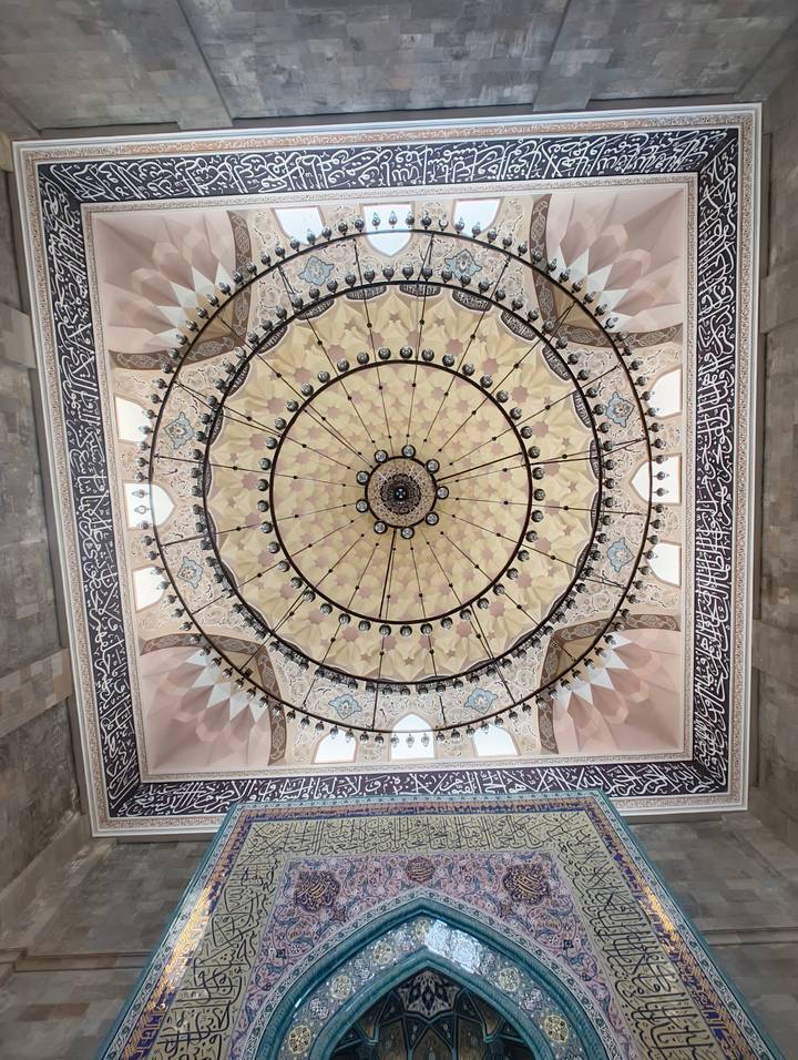 Symmetrical ornate mosque ceiling with concentric floral patterns, chandeliers and Arabic calligraphy.