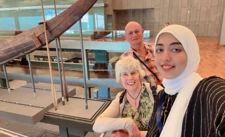 Smiling tourists posing for a selfie inside a modern museum with large ancient wooden boat on display.