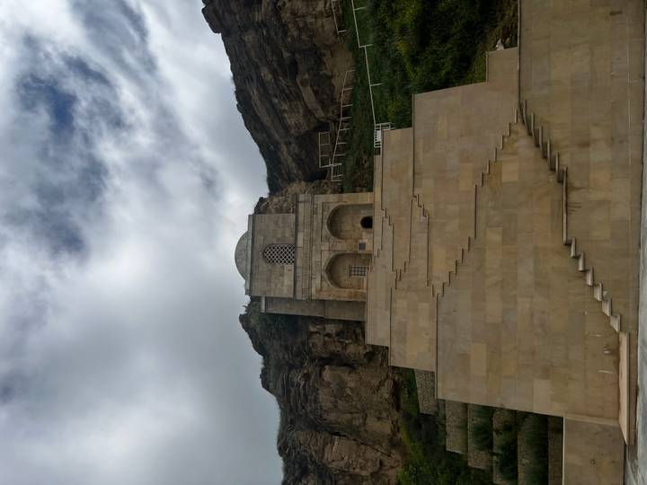 Tall beige stone mausoleum built into a cliff face viewed from below under cloudy skies.