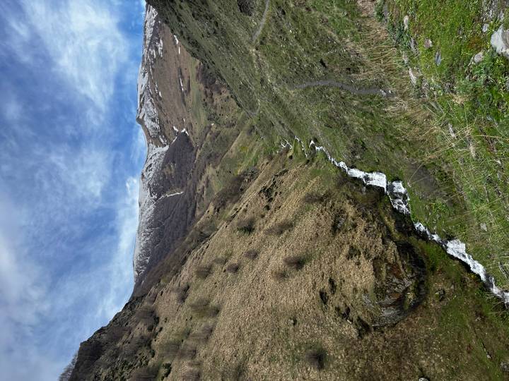 Mountain valley with melting snow patches, a small cascading stream, and dramatic cloudy sky overhead.