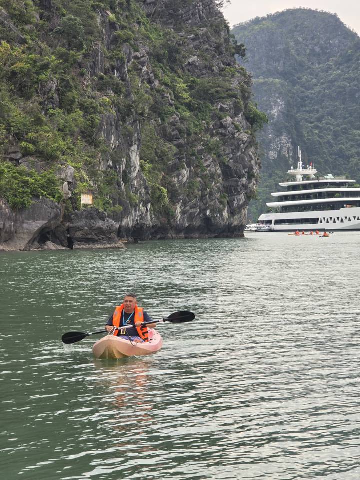 Man in kayak paddling through emerald waters with towering limestone cliffs and cruise ship behind.