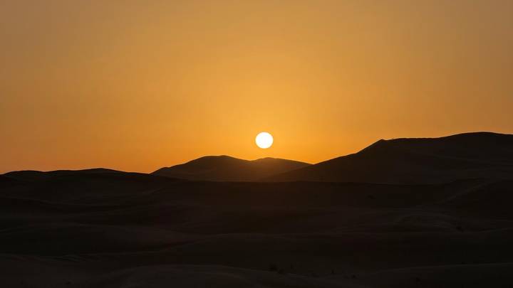 Golden sun setting behind rolling desert dunes casting deep shadows.