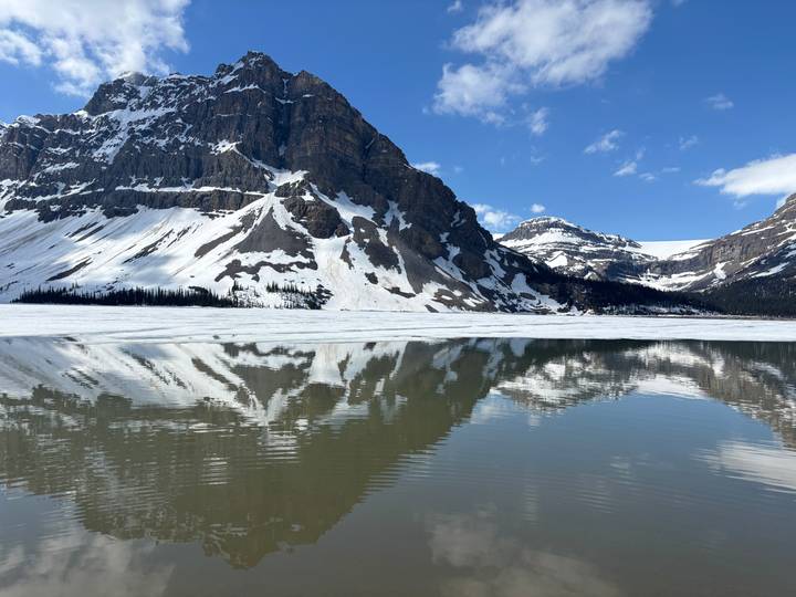 Snow-clad rocky peak reflecting in a partially frozen turquoise lake under a bright blue sky.