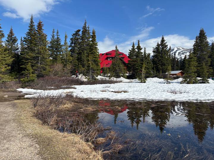 Bright red alpine lodge nestled among tall pine trees, snow patches and a still reflective pond.