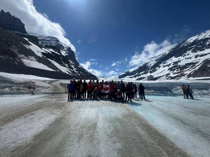 Large tour group standing on a bright blue glacier surrounded by steep snowy mountains.