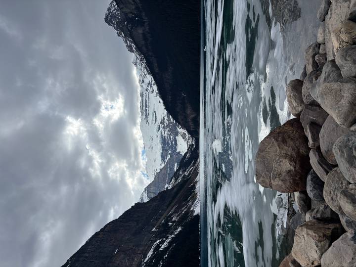 Part-frozen Lake Louise lined with boulders, ringed by snowy peaks under a moody cloudy sky.