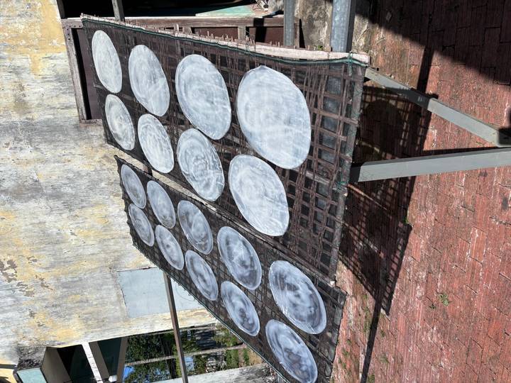 Round rice paper sheets dry in the sun on a mesh rack against a weathered stucco wall.