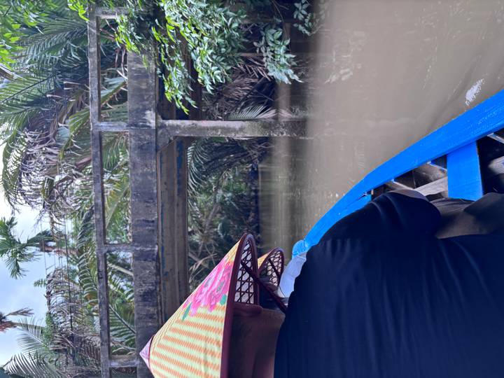 A traveller wearing a conical hat paddles a blue sampan beneath a low concrete bridge through muddy jungle waters.