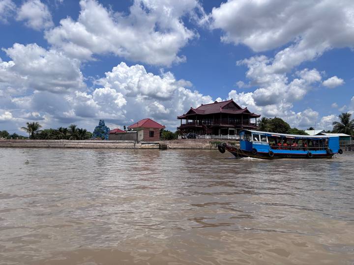 A blue tourist boat cruises along a wide brown river with wooden stilt houses and a cloud-filled sky above.