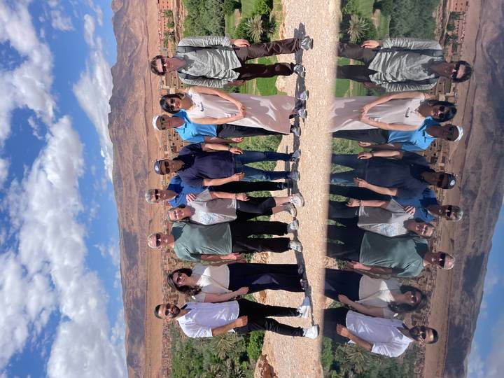 Tour group posing with arid Moroccan mountains and desert village in the background, creative mirror reflection doubling the group below.