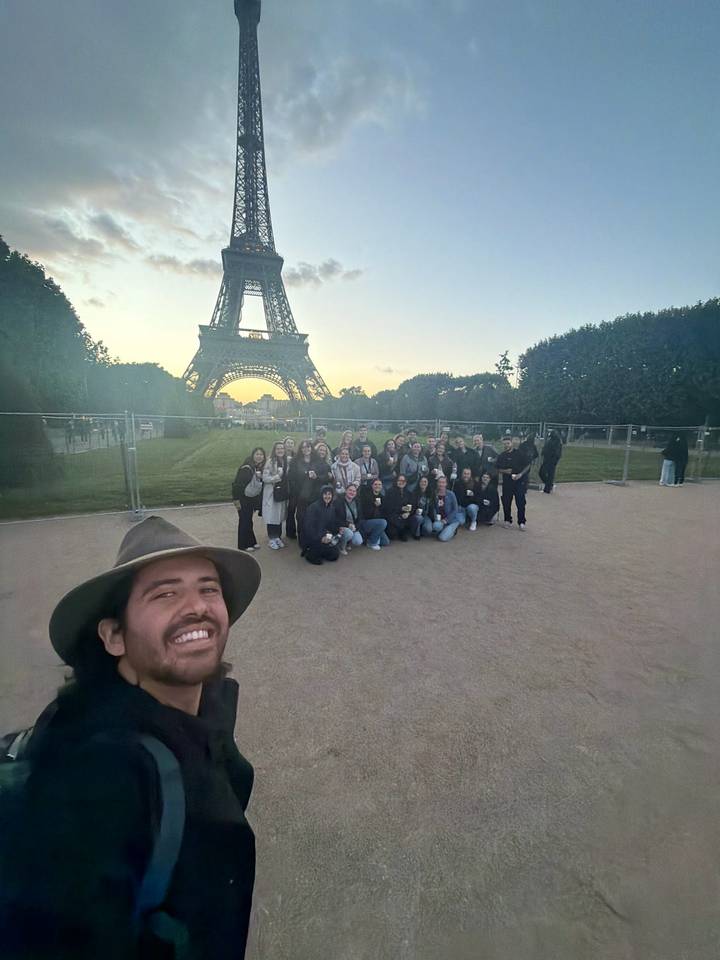 Large tour group smiling for a selfie in front of the Eiffel Tower at dusk.
