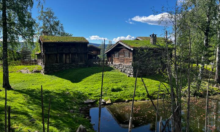 Traditional Norwegian turf-roof farm buildings beside a pond under a clear sky near Lillehammer.
