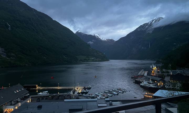 Evening view down Geirangerfjord with snow-capped peaks rising above a small harbor’s lights.