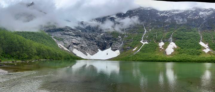 Serene alpine lake with melting snow patches beneath misty peaks reflected in green water.