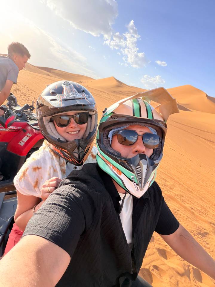 Selfie of two people wearing helmets on quad bike against orange desert dunes
