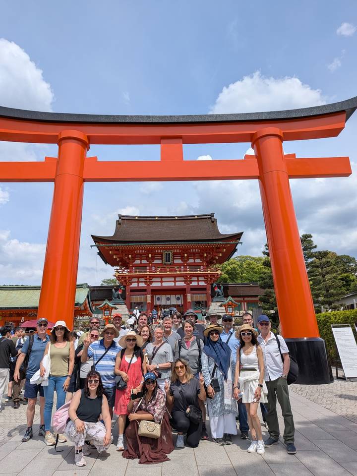 Tour group gathered beneath a massive vermilion torii gate with the ornate entrance of Fushimi Inari Shrine behind them in Kyoto.