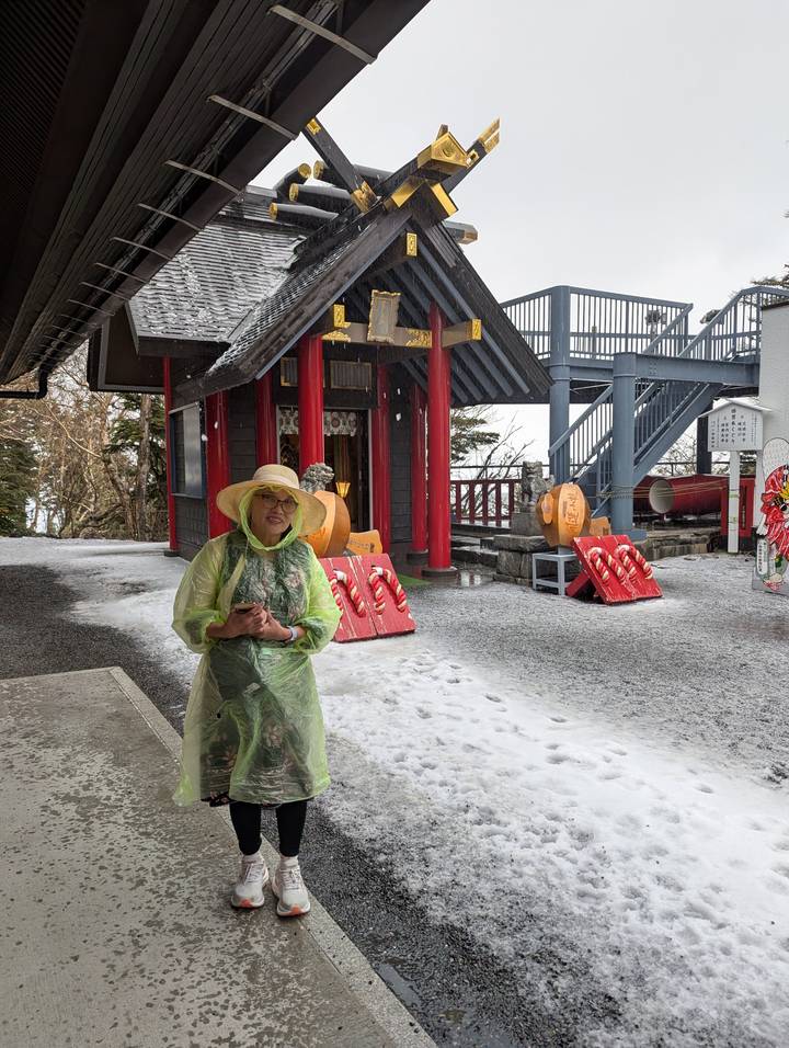 Woman wearing a rain poncho and sun hat stands in front of a small red Shinto shrine building dusted with fresh snow on an overcast day.