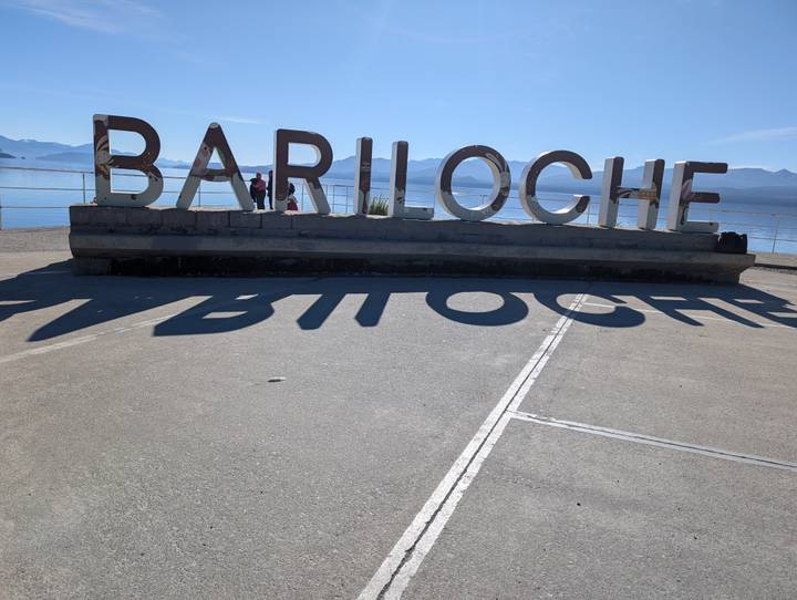 Large BARILOCHE letters cast long shadows beside a sparkling Patagonian lake backed by distant mountain ranges.