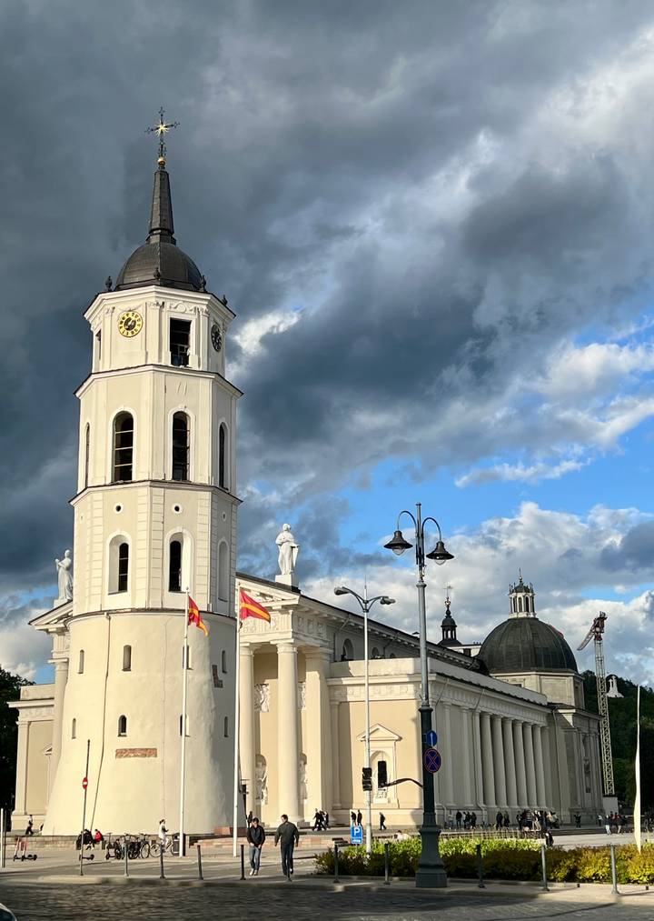 Tall white bell tower with statues and national flags under a dramatic cloud-filled sky.