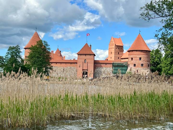 Picturesque red-brick Trakai Castle with towers rising above reeds against a blue sky.