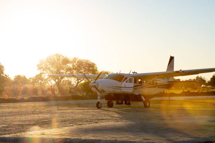 Light aircraft parked on sandy runway at sunrise with warm lens flare and silhouettes of trees.