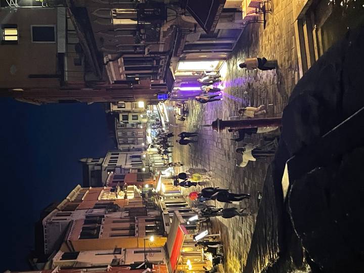 A lively Venetian street at night is lit by shopfronts and cafés as people stroll along wet cobblestones.