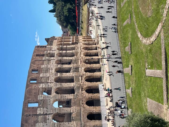 The Colosseum’s ancient arches tower over visitors walking the sunny grounds of Rome’s iconic arena.