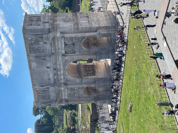 The Arch of Constantine stands near the Colosseum as tourists gather below on a bright day.