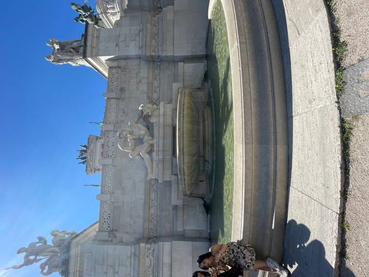 A sculpted fountain with reclining figure sits in front of the Vittoriano monument under clear blue sky.