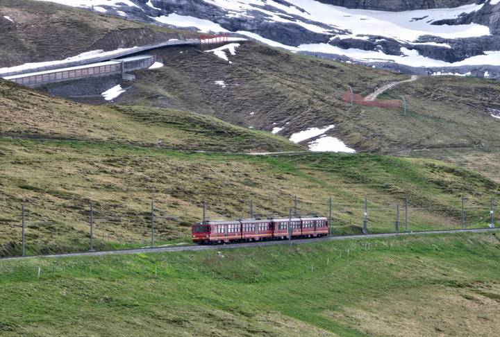 Red cog railway train climbing green alpine slopes with patches of late-season snow