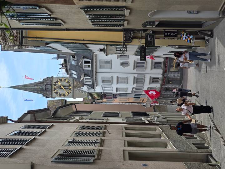 Steep cobbled Zurich lane with clock tower, Swiss flags and pedestrians between shuttered houses