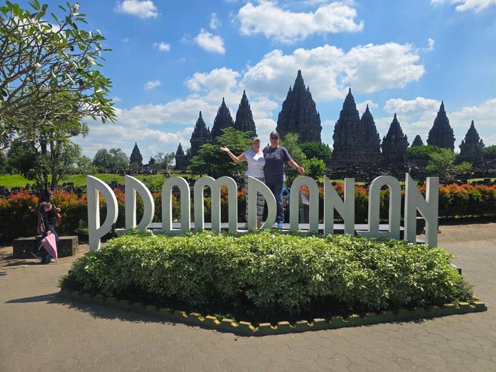Travellers posing atop landscaped sign spelling Prambanan with temple spires in background