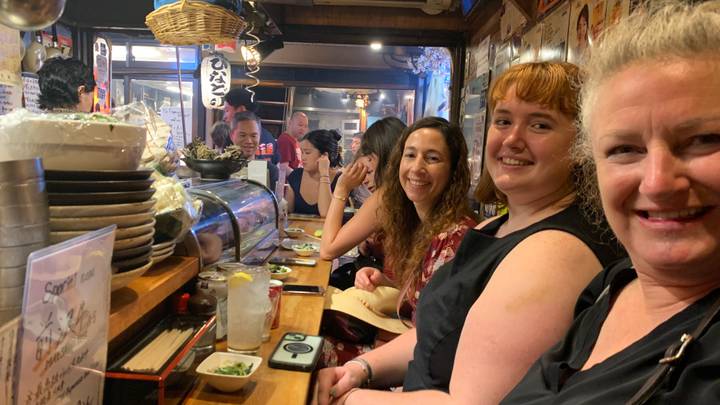 Lively diners seated shoulder-to-shoulder at a small Japanese izakaya counter filled with dishes and lanterns