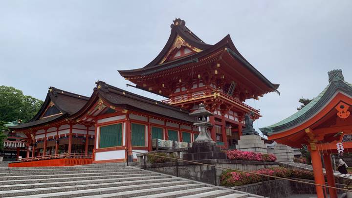 Grand red-and-white main shrine buildings of Fushimi Inari rise above stone steps under a gray sky