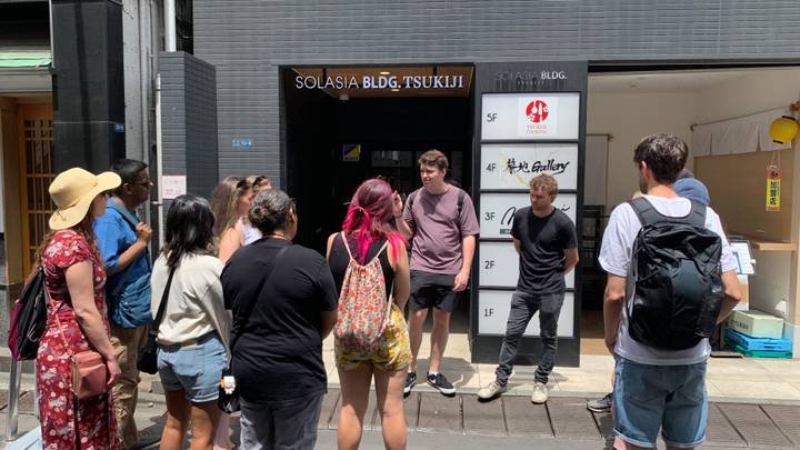 Guide speaks to a cluster of travelers outside a modern building labeled Solasia Bldg Tsukiji in Tokyo