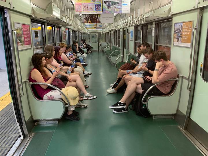 Passengers seated along both sides of a green-floored Japanese train carriage, many on phones