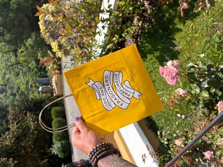 Close-up of a yellow Fragonard shopping bag held outdoors near blooming flowers.