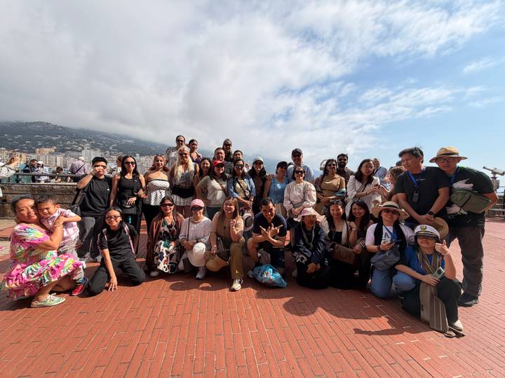 Large tour group posing on a terrace overlooking Monaco with coastal hills in background.