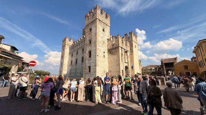 Crowd of tourists gathered outside a medieval stone castle tower on a sunny day.