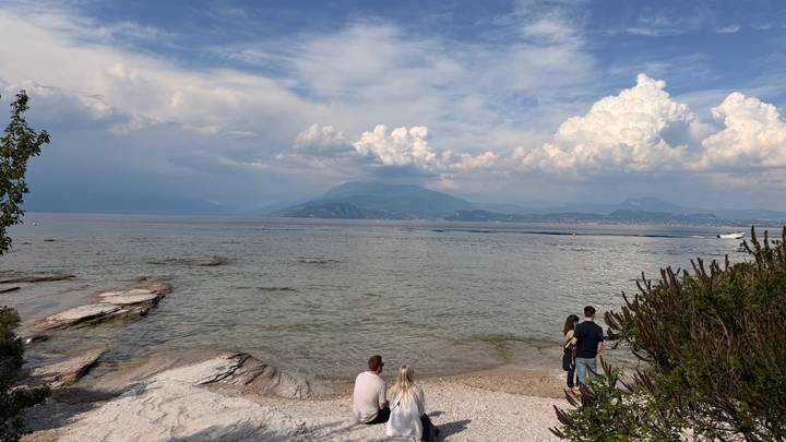 Small group relaxing by a rocky lakeshore with billowing clouds over distant peaks.