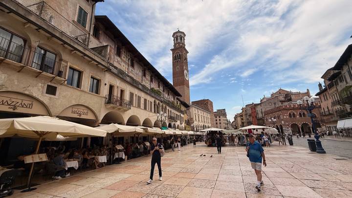 Lively Italian piazza lined with cafés and umbrellas, dominated by a tall clock tower.